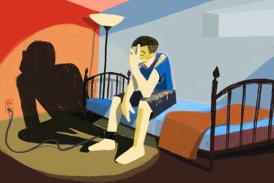 A teenager sitting on their bed while charging an ankle monitoring bracelet, and a surreal aggressive spotlight shines onto them.
