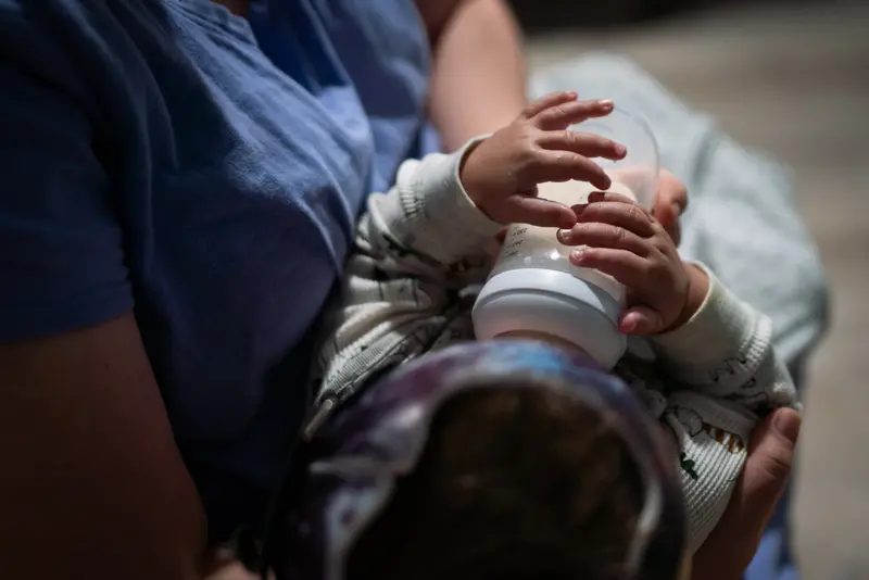 An adult’s arms hold a small baby who’s drinking from a bottle.